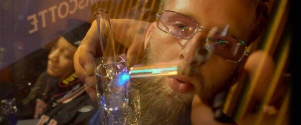 Fragile glass pipe with colorful lighting reflections, person in glasses smoking marijuana at a cannabis event, vibrant and close-up shot capturing the smoking process, fantastic for cannabis and smoking related SEO.