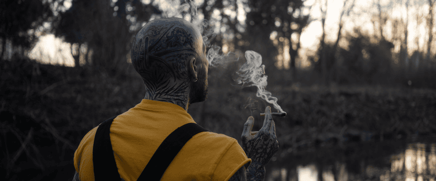 Man with tattoos smoking outdoors during sunset, featuring nature background and a relaxed atmosphere.