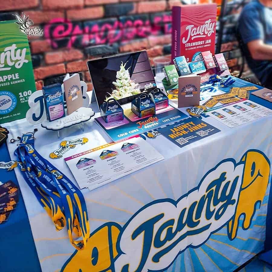 Brightly colored promotional display table for Taunty gummies and products at AudioKush event. Featuring various gummies, branded merchandise, and informational flyers for cannabis-infused edible products.