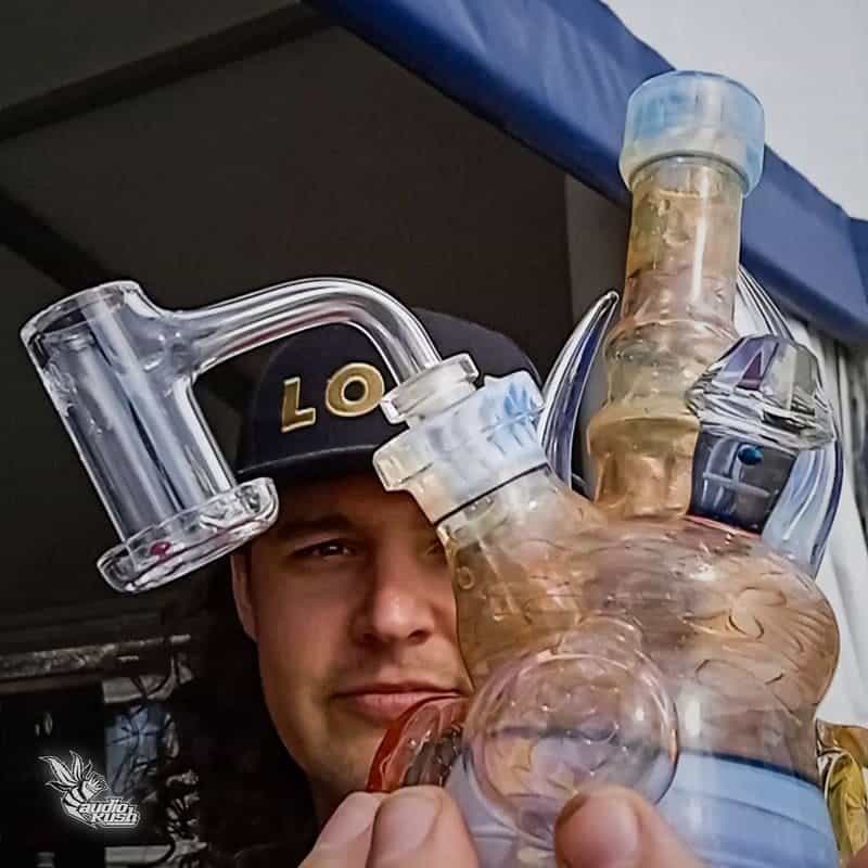 Colorful glass bong and a person wearing a hat with "LO" letters, ready for a smoke session, at an outdoor event or festival.