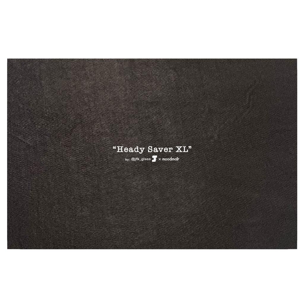 Dark textured surface with white text displaying "Heady Saver XL" by @jfk_glass x moodmat, showcasing a premium black moodmat for smoking accessories.