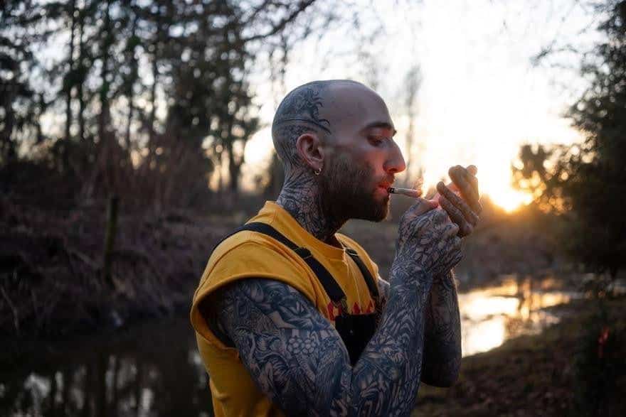 Vape smoker with tattoos outdoors during sunset, enjoying a relaxing moment by the river - alternative lifestyle and cannabis culture.