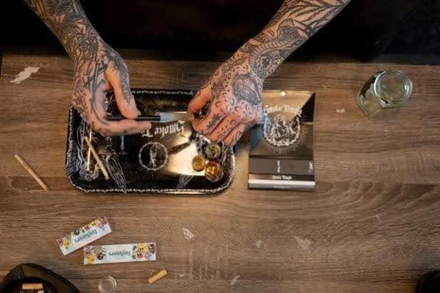 Tattooed hands handling a grinder with cannabis and smoking accessories on wooden table, cannabis cannabis, recreational drug use, cannabis grinder, drug paraphernalia.